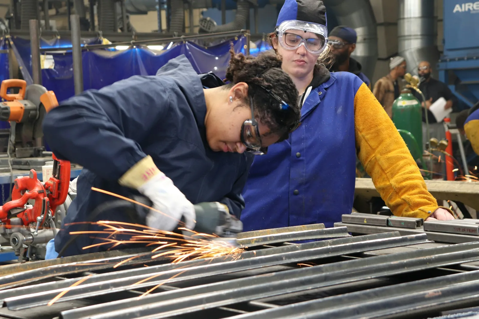 A welder grinds a steel frame in a workshop while an instructor in a face shield watches beside him.